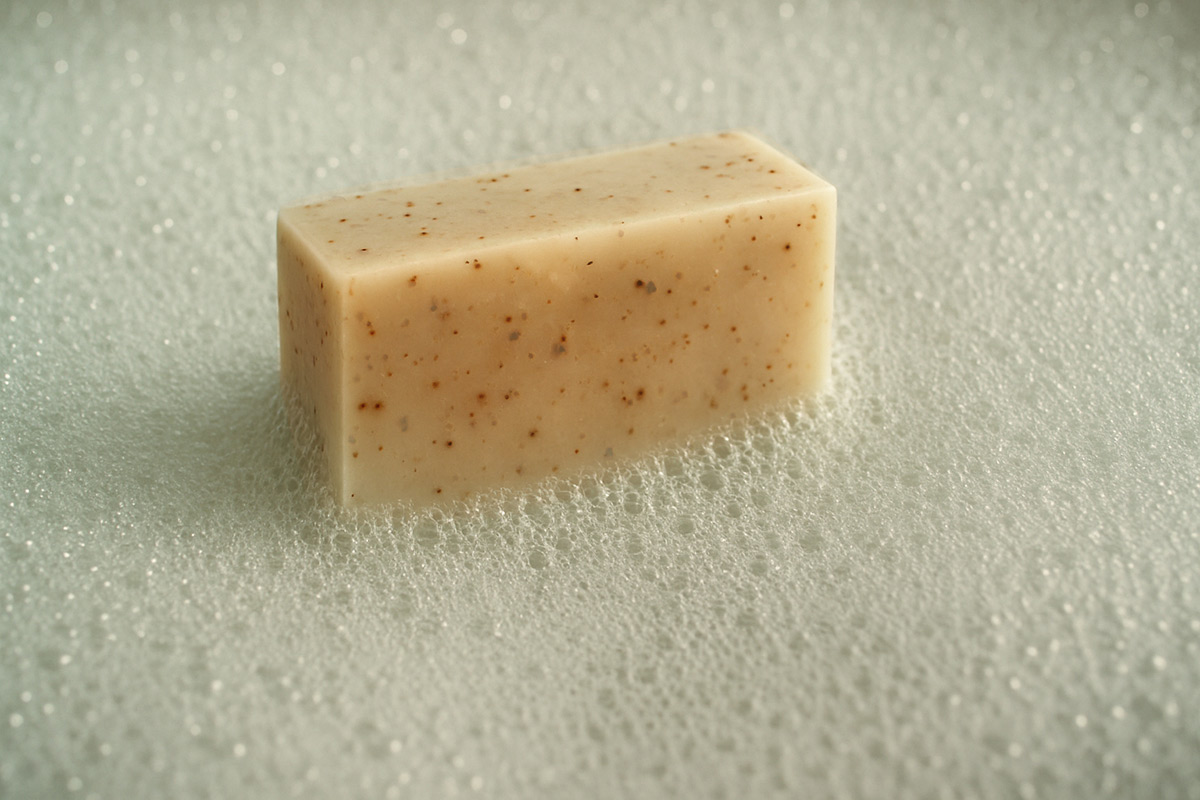 A handcrafted exfoliating soap bar with visible flecks sits delicately in a soft bed of pale green-beige bubbles. Captured in natural light, the scene evokes a serene and clean atmosphere.